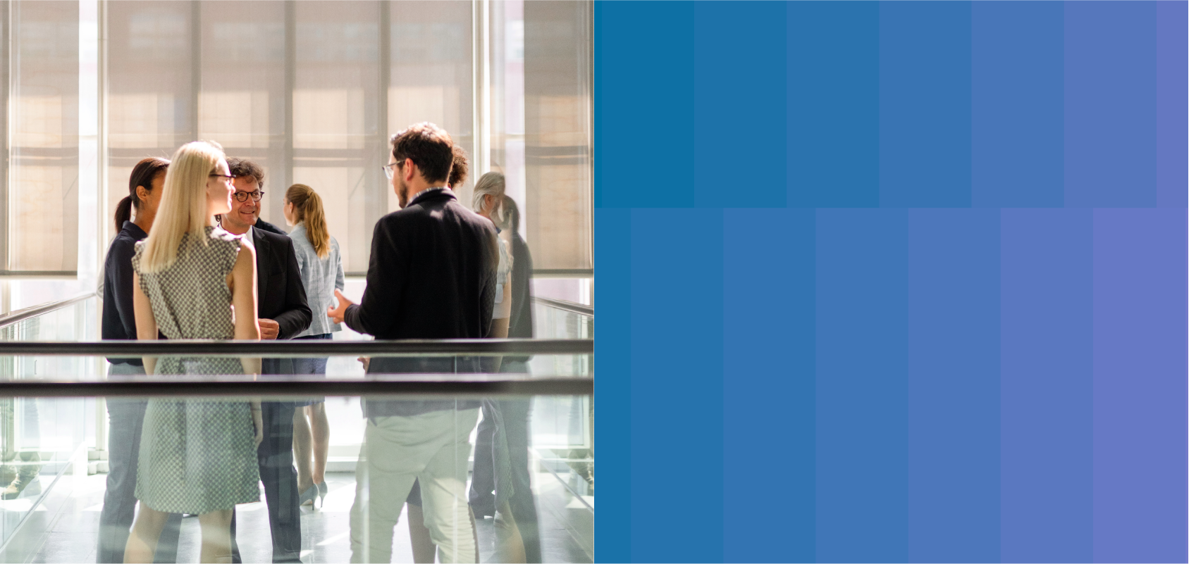 photo, employees chat at a gathering in a large office hallway. They are standing near waist-high glass guard rails with brown banisters. A large building can be seen through the open window nearby.