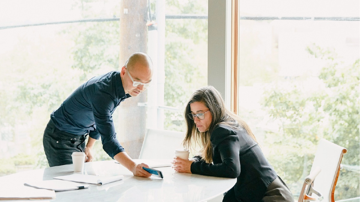 Two colleagues review information on a tablet at a table in a bright office.